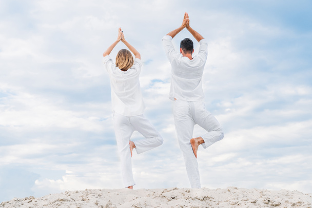 rear view of couple in white clothes practicing yoga in tree pose (Vrksasana) on sandy dune - Fotó, kép