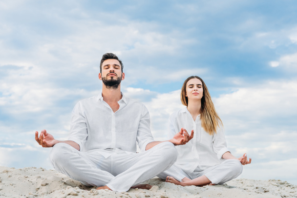 calm young couple meditating while sitting on sandy dune in lotus pose (padmasana) - Foto, immagini