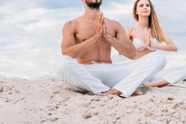 cropped shot of young couple meditating while sitting on sandy dune in lotus pose (padmasana) - 写真・画像