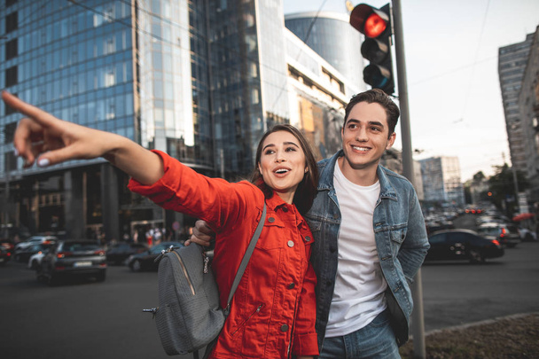Waist up portrait of delighted girl pointing finger on street. Happy boyfriend is standing near her looking with content - Fotó, kép