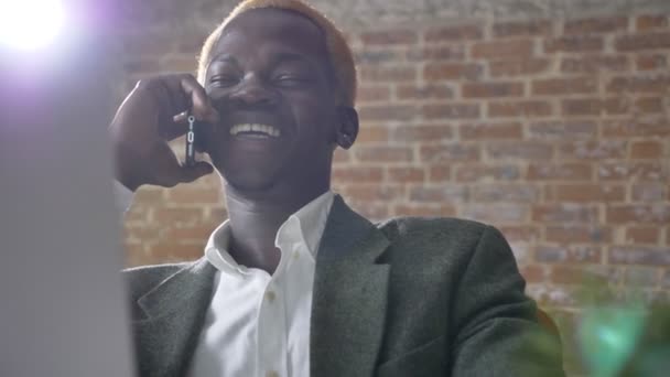 Young african happy blonde businessman talking on phone and sitting in modern office, laughing and smiling - Materiaali, video