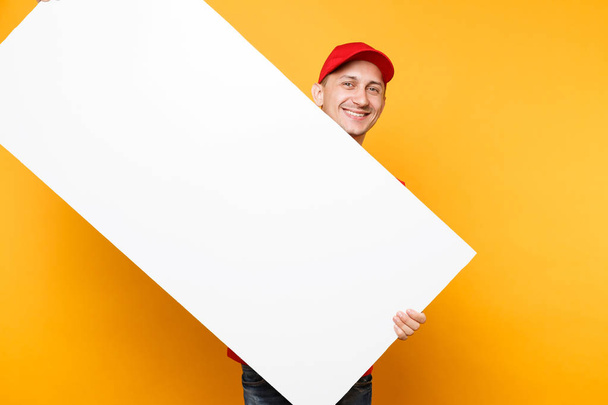 Чоловік в червоній формі ізольований на жовтому помаранчевому фоні. Кур'єр чоловічої статі в шапці, футболка, що тримає великий білий порожній рекламний щит з копією, місце для текстового зображення. Концепція сервісу
 - Фото, зображення