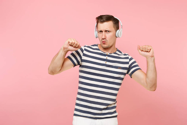 Портрет улыбающегося молодого человека в полосатой футболке, слушающего музыку в белых беспроводных наушниках, наслаждающегося изолированным на модном пастельно-розовом фоне. Люди искренние эмоции
 - Фото, изображение
