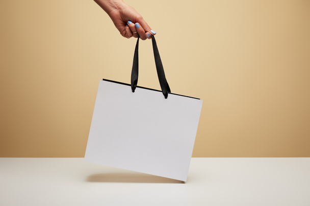 cropped image of woman holding white shopping bag above white table isolated on beige - Photo, Image