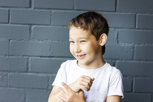 Lindo niño sonriente de 8 años confundido en fondo de pared de ladrillo gris
 - Foto, Imagen