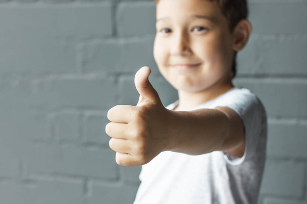 Cute smiling boy 8 years old showing thumbs up on background of grey brick wall - Fotoğraf, Görsel