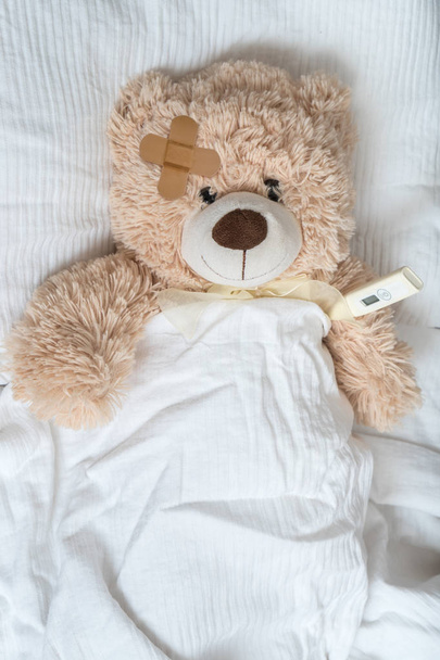 Urso de pelúcia doente com gesso e termômetro está deitado na cama
. - Foto, Imagem