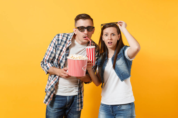 Young shocked couple woman and man in 3d glasses watching movie film on date holding bucket of popcorn drinking soda or cola from plastic cup isolated on yellow background. Emotion in cinema concept - Foto, Bild