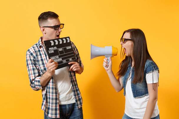 Young irritated couple woman man in 3d glasses watching movie film on date hold classic black film making clapperboard screaming on megaphone isolated on yellow background. Emotions in cinema concept - Fotó, kép