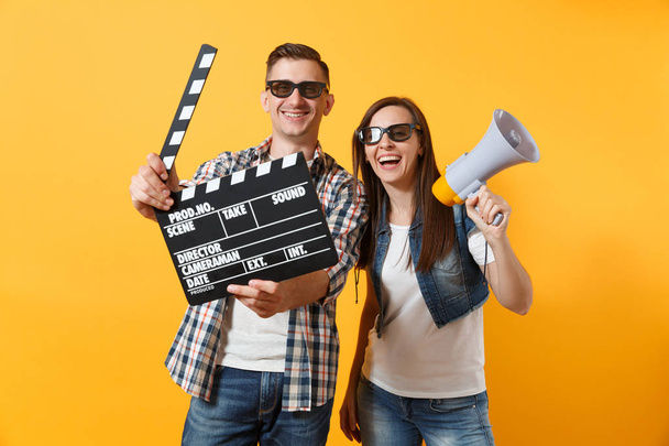 Молодой счастливой улыбчивой паре мужчина в 3D очках смотрит фильм на свидании, держа классический чёрный фильм, делающий клапперборд и мегафон изолированным на жёлтом фоне. Эмоции в концепции кино
 - Фото, изображение