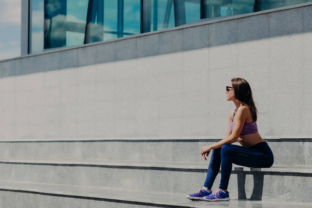 Horizontal shot of beautiful sporty woman in casual clothes, sits on stairs, has rest after gymnastics, poses outdoor. Female athlete in sneakers trains in open air. People, active lifestyle concept - Фото, изображение