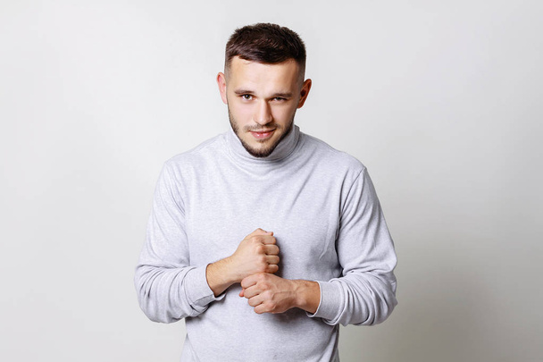 Close up portrait of a young man smirking. Handsome in white turtleneck beats his fist on the fist standing on grey background copy space - Photo, Image