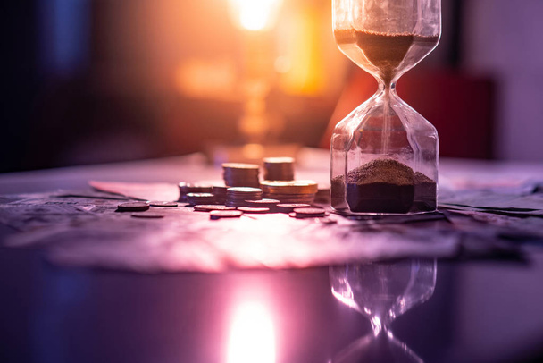 Sand running through the shape of hourglass on table with banknotes and coins of international currency. Time investment and retirement saving. Urgency countdown timer for business deadline concept - Photo, Image