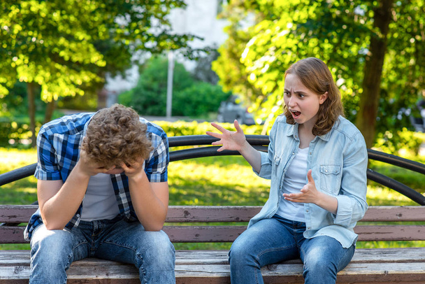 The girl screams at young man. Summer in park on a bench. The guy is crying and sad, clasping his head in his hands. Scandal in family. The concept of aggression and the problem in relationship. - Photo, Image
