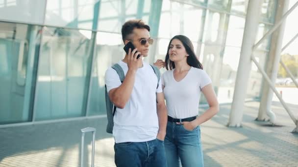 Attractive young couple standing near the airport. A man calls a taxi or call the delivery service. Communication, technology, smartphone concept - Materiaali, video