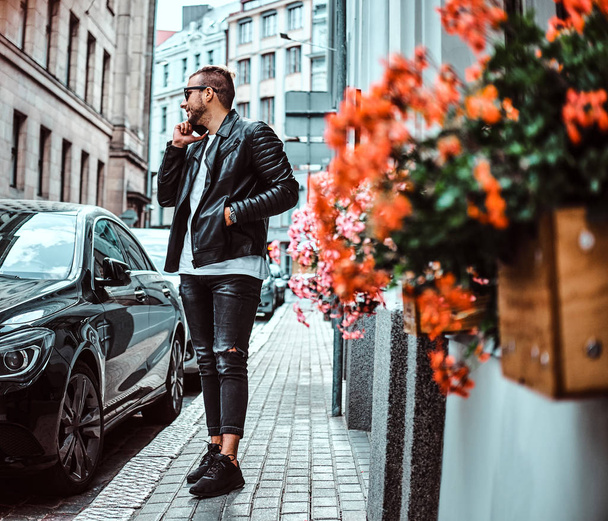 Modny facet w okulary ubrany w czarną skórzaną kurtkę i dżinsy rozmowy przez smartfona, stojąc w pobliżu luksusowy samochód na starej ulicy Europy. - Zdjęcie, obraz