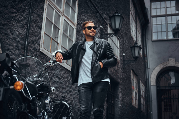 Fashionable biker in sunglasses dressed in a black leather jacket and jeans leaning on his retro motorcycle on old Europe street. - 写真・画像