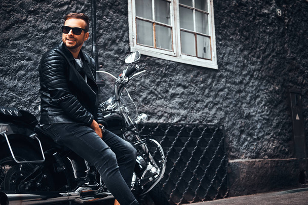 Muodikas nuori moottoripyöräilijä aurinkolaseissa pukeutunut musta nahkatakki ja farkut istuu hänen retro moottoripyörä vanhan Euroopan kadulla
. - Valokuva, kuva