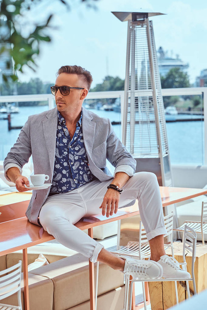Pewny siebie człowieka sukcesu z stylowe włosy, ubrany w eleganckie nowoczesne ubrania posiada kawę siedząc na stole w kawiarni na świeżym powietrzu na tle nabrzeża. - Zdjęcie, obraz