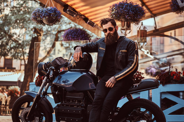 Портрет стильного модного байкера в солнцезащитных очках, одетый в черную кожаную куртку, сидящего на своем ретро-мотоцикле на заказ рядом с террасой кафе
. - Фото, изображение