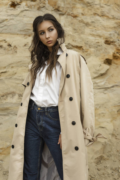 Beautiful girl wearing blue jeans, a white blouse and a beige raincoat posing in a sandy quarry against the backdrop of a sand wall. Healthy clean skin. Commercial, fashionable and advertising photo - Valokuva, kuva