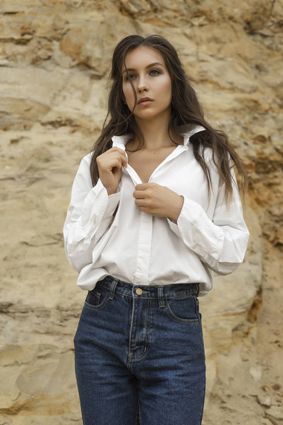 Beautiful girl wearing blue jeans, a white blouse unfastens her blouse in a sandy quarry against the backdrop of a sand wall. Healthy clean skin. Commercial, fashionable and advertising photo - Foto, immagini