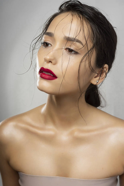 Beautiful brunette girl fashion model with naked shoulders and wet hair on a gray background. Nude make-up, red lips. Healthy clean skin. Advertising and commercial design. - Φωτογραφία, εικόνα