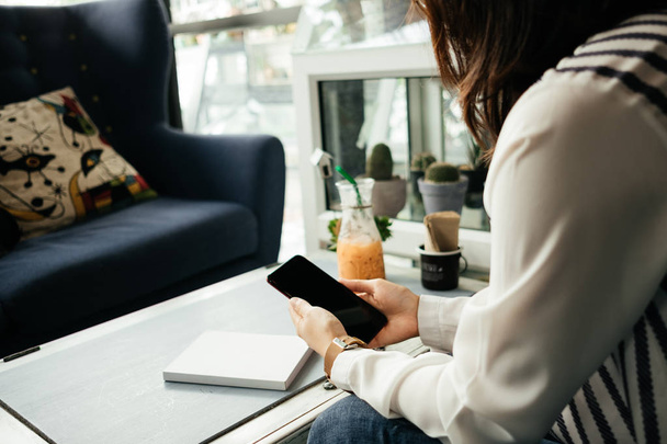 молода бізнес-леді, сидячи на робочому місці і використовуючи мобільний телефон, має блокнот і напій на фоні столу
 - Фото, зображення