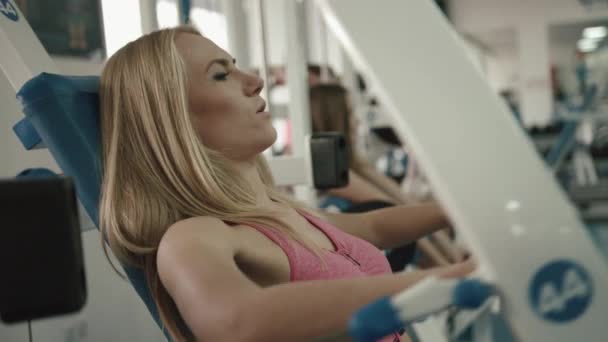 Attractive athletic blonde woman working out at gym. She is doing exercises for arms on trainor. 4k footage. - Imágenes, Vídeo