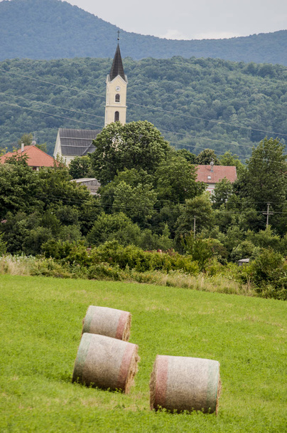 Croatie, 26 / 06 / 2018 : Foin jaune avec prairies verdoyantes sur des terres cultivées à la campagne et l'église paroissiale du petit village de Drenik Grad, dans la région des lacs de Plitvice
  - Photo, image