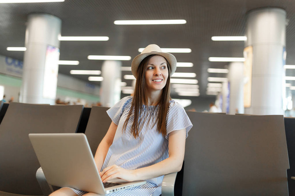Giovane turista viaggiatore sorridente donna in cappello che lavora sul computer portatile, guardando da parte durante l'attesa nella hall dell'aeroporto internazionale. Passeggeri che viaggiano all'estero durante il fine settimana. Concetto di volo aereo
 - Foto, immagini