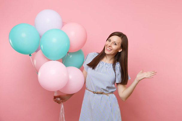Portrait of smiling young happy woman wearing blue dress holding colorful air balloons pointing hand aside on copy space isolated on bright trending pink background. Birthday holiday party concept - Фото, изображение