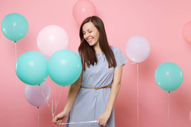 Portrait of attractive laughing young woman with closed eyes in blue dress holding colorful air balloons on bright trending pink background. Birthday holiday party, people sincere emotions concept - Zdjęcie, obraz