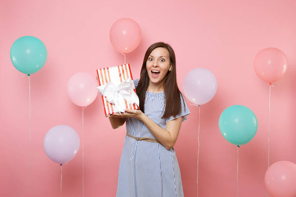 Portret van verrast, verbaasd jonge vrouw in blauwe jurk met rode doos met cadeau op pastel roze achtergrond met kleurrijke lucht ballonnen aanwezig. Vakantie verjaardagsfeestje, mensen oprechte emoties concept - Foto, afbeelding