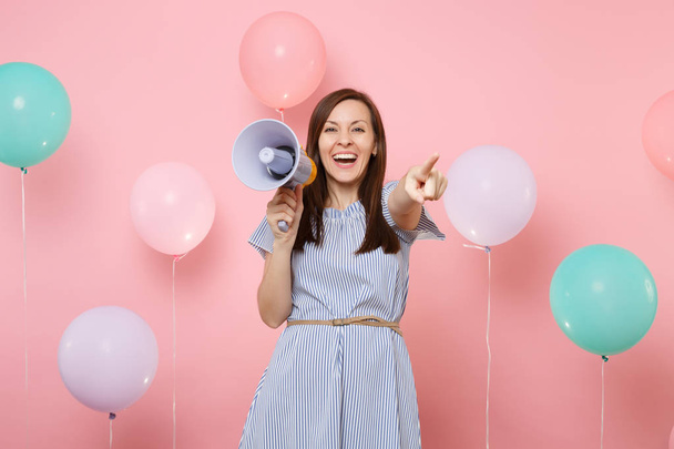 Portrait of laughing young pretty woman in blue dress holding megaphone pointing index finger on camera on pink background with colorful air baloons. Birthday holiday party, people sincere emotions - Foto, Imagen
