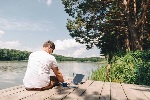 człowiek pracuje na laptopie siedząc drewnianych dok. nogi w wodzie rzeki - Zdjęcie, obraz