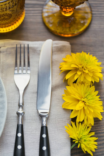Rustic table setting with linen napkin, cutlery, ceramic plates, yellow glasses and yellow flowers on dark wooden table. Holiday table decoration. Romantic dinner. - Foto, Imagem