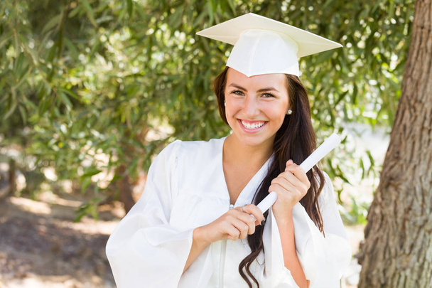 Atractiva chica de raza mixta celebrando la graduación en el exterior con gorra y vestido con diploma en la mano
. - Foto, Imagen