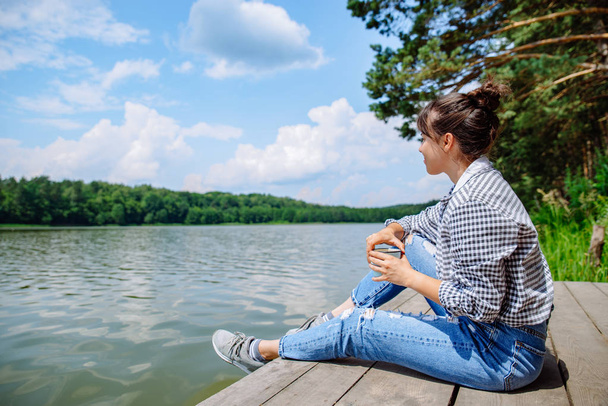 молодая взрослая женщина, сидящая на деревянном причале, пьет кофе и смотрит на озеро
 - Фото, изображение