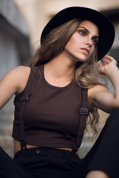 Portrait of a cute young white Caucasian girl in a black stylish hat and a short brown top. Fashion portrait of a sexy girl at sunset. - Foto, immagini