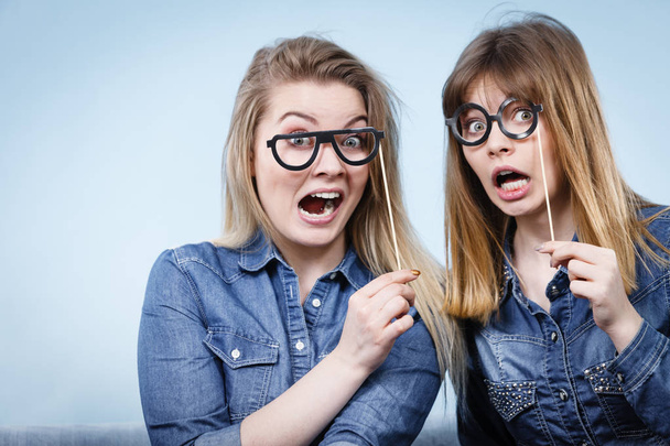 Two happy women holding fake eyeglasses on stick having fun fooling around wearing jeans shirts. Photo and carnival funny accessories concept. - 写真・画像
