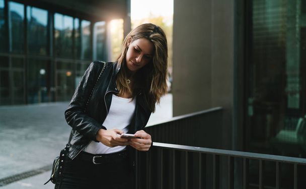 Hipster girl wearing black leather jacket and black jeans reading emails on a mobile phone while standing outdoors on a blurred urban background. Student girl chatting online by a smartphone outdoors - Foto, immagini