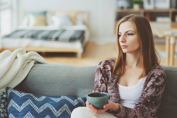indoor lifestyle portrait of young woman relaxing at home with cup of hot tea or coffee, sitting on couch. Modern scandinavian apartment interior - Fotoğraf, Görsel