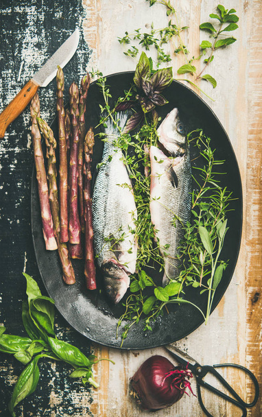 raw uncooked sea bass fish with fresh herbs and vegetables on dark plate over rustic wooden painted background - Foto, Bild