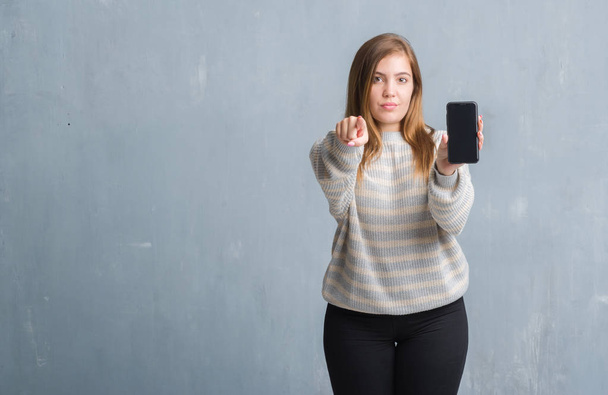 Jovem mulher adulta sobre parede grunge cinza mostrando tela em branco do smartphone apontando com o dedo para a câmera e para você, sinal de mão, gesto positivo e confiante da frente
 - Foto, Imagem