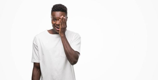Giovane uomo afroamericano indossa una t-shirt bianca che copre un occhio con la mano con un sorriso fiducioso sul viso e l'emozione sorpresa
. - Foto, immagini