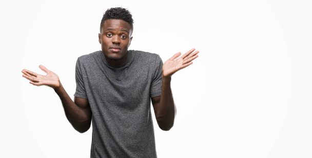 Jonge Afro-Amerikaanse man met grijs t-shirt clueless en verward expressie met armen en handen aan de orde gesteld. Twijfel concept. - Foto, afbeelding
