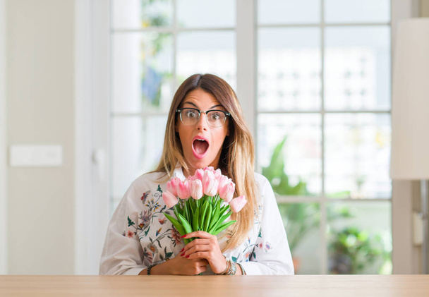 Giovane donna a casa in possesso di tulipani rosa fiori spaventati in stato di shock con un volto a sorpresa, spaventati ed eccitati con espressione di paura
 - Foto, immagini