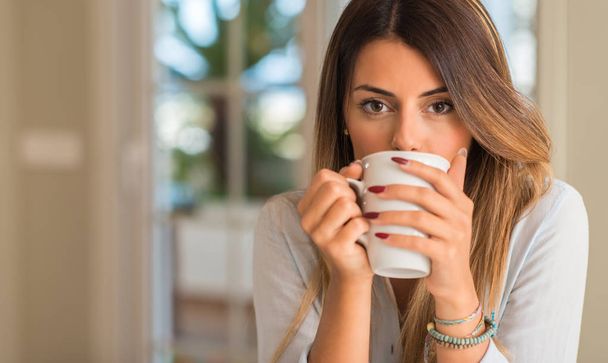 美しい若い女性がカメラを見て手でカップを押しながらコーヒーを飲みます。家での朝食 - 写真・画像