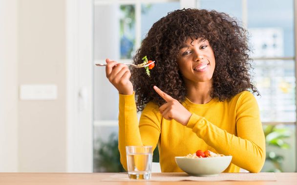 Mujer afroamericana comiendo ensalada de pasta en casa muy feliz señalando con la mano y el dedo hacia un lado
 - Foto, imagen